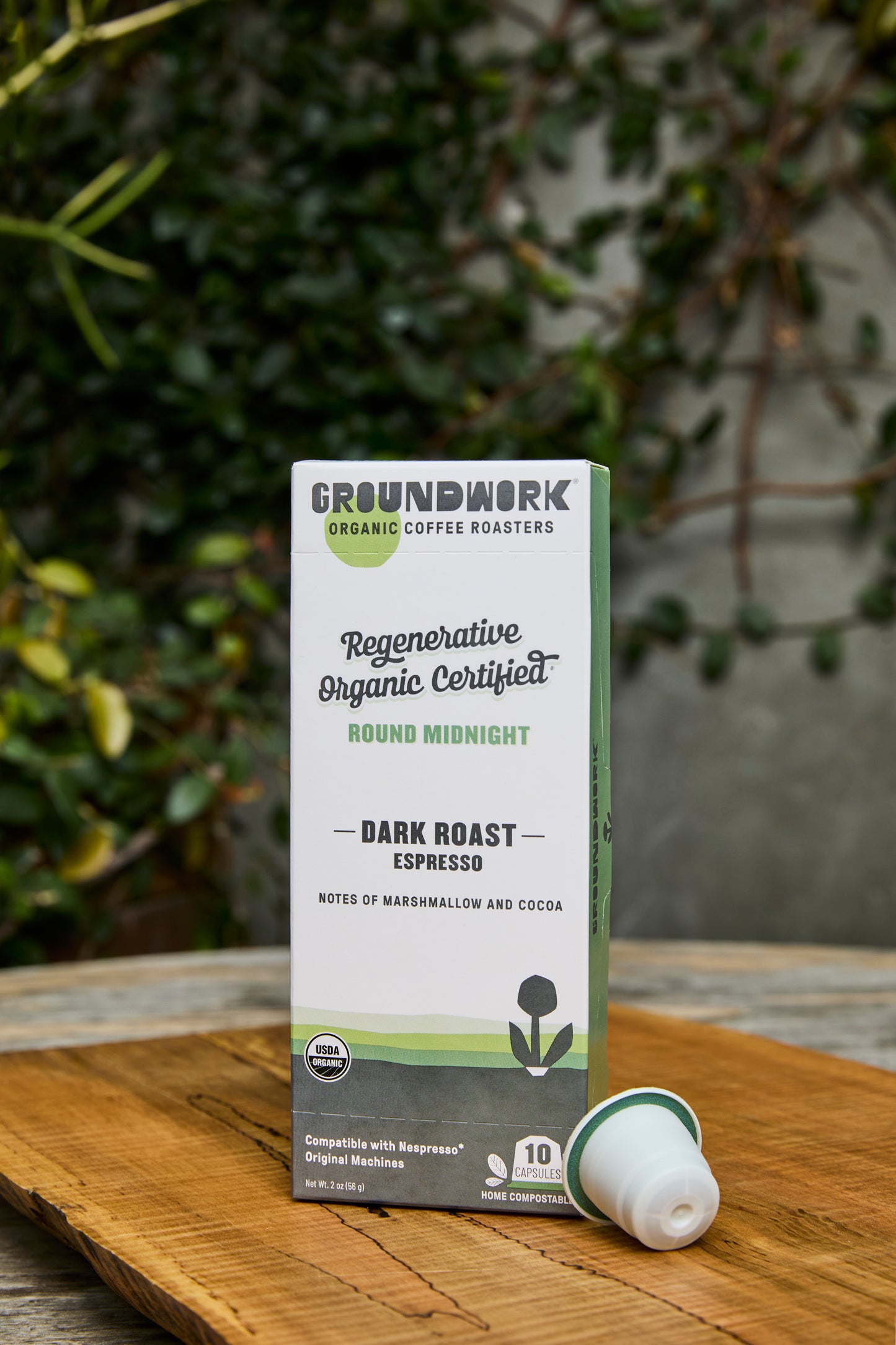 A box of Groundwork Coffee Regenerative Organic Certified® Round Midnight - Nespresso® pods sits outdoors on wood with greenery behind it; one pod is placed next to the box.