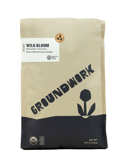 A 5 lb beige and black bag of Groundwork Coffee Regenerative Organic Certified® Wild Bloom medium roast whole bean, featuring milk chocolate and apple notes with a flower graphic.