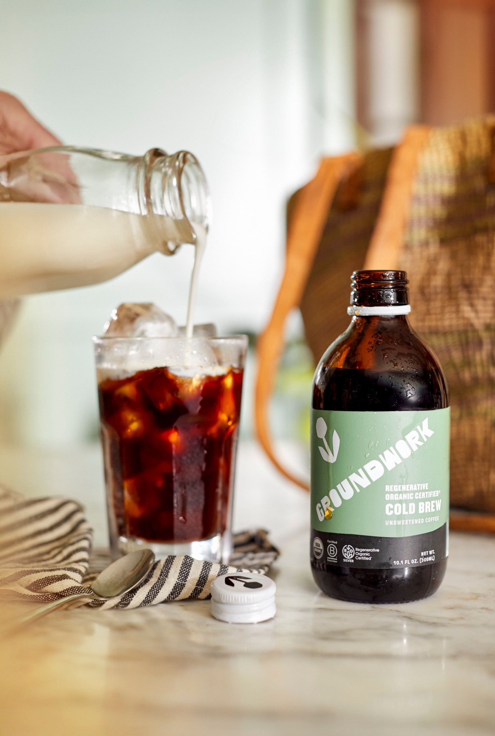A hand pours milk into iced coffee next to a Regenerative Organic Certified® Cold Brew by Groundwork Coffee Co on a marble countertop.