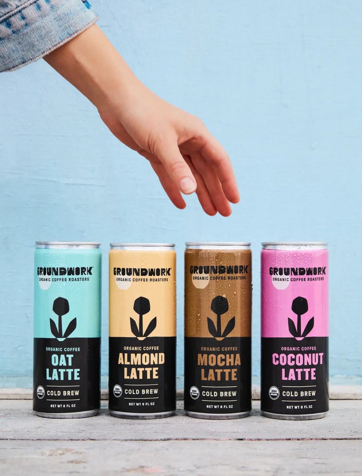 A hand reaches for one of four Groundwork Coffee Organic Cold Brew Latte cans—Oat, Almond, Mocha, and Coconut—lined up against a light blue background.