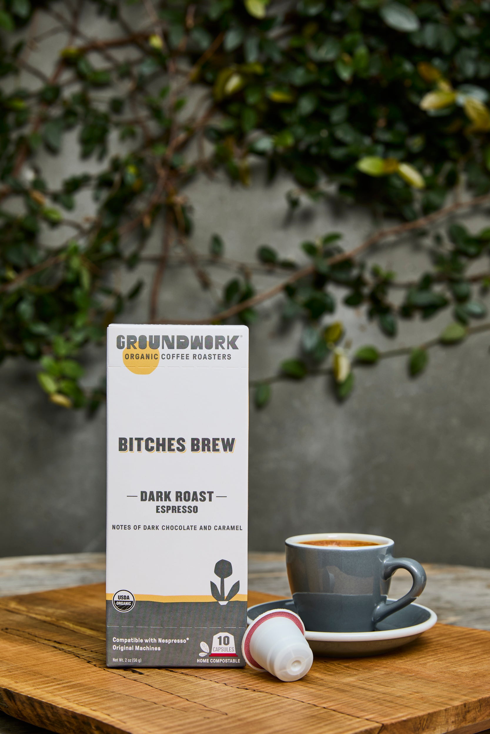 A box of Groundwork Coffee Bitches Brew - Nespresso® pods sits on a wooden table beside a grey cup, with leafy plants in the blurred background.