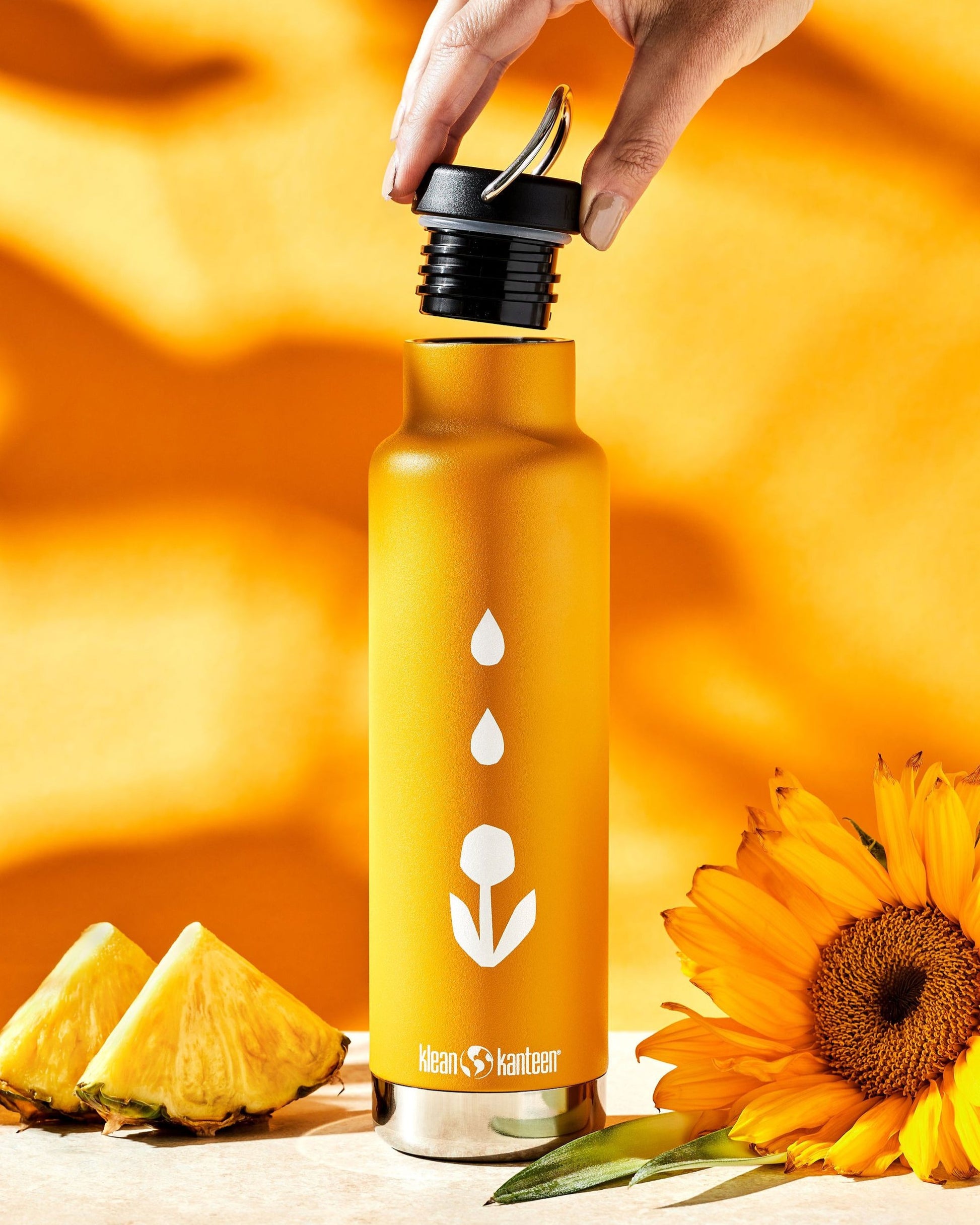 A hand opens the cap of a yellow Klean Kanteen 20oz Insulated Water Bottle by Groundwork Coffee, with pineapple slices and a sunflower on a matching yellow background.
