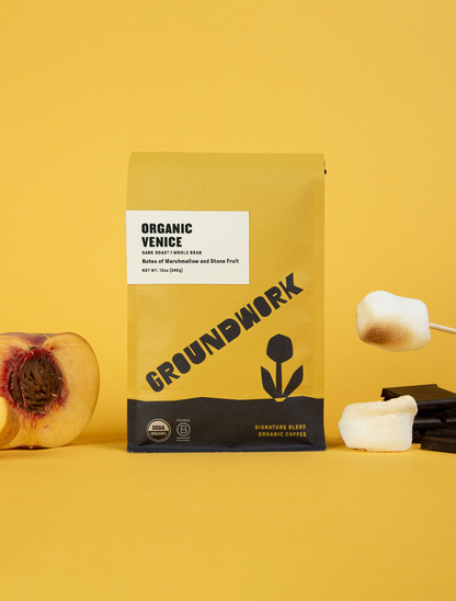 A yellow bag of Groundwork Coffees Venice sits on a yellow surface, beside stone fruit, toasted marshmallows, and chocolate.
