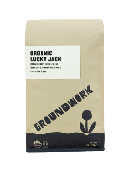 A brown bag of Groundwork Coffees Lucky Jack whole bean coffee, a medium roast with caramel and citrus notes, features a flower graphic and blend certifications.