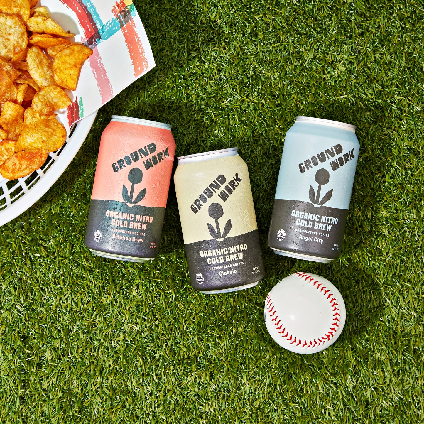 Three cans of Groundwork Coffee’s Organic Angel City Nitro Cold Brew sit on green grass next to a baseball and a plate of potato chips.