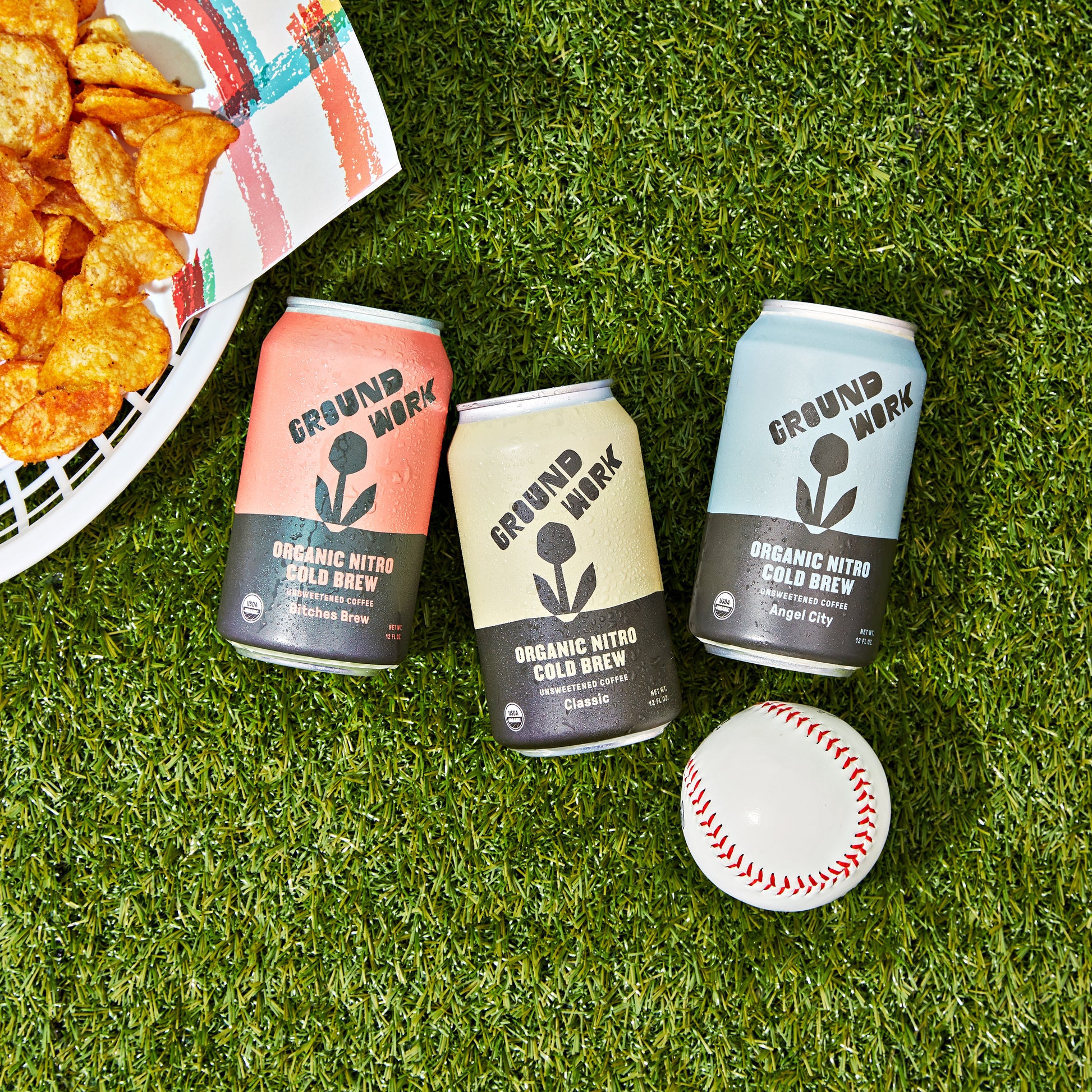 Three cans of Groundwork Coffee’s Organic Angel City Nitro Cold Brew sit on green grass next to a baseball and a plate of potato chips.