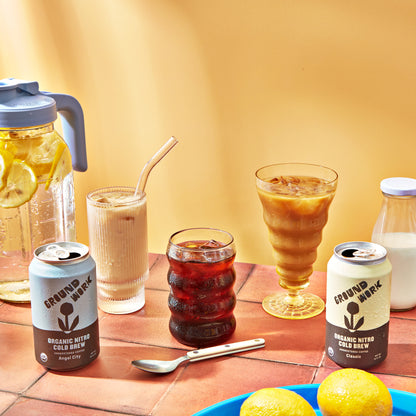 A table with cold drinks, featuring two cans of Groundwork Coffee Organic Angel City Nitro Cold Brew, iced coffee in glasses, lemon water in a pitcher, milk, and fresh lemons.